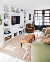 Sofa with scatter cushions and ottoman opposite wall with masonry shelving and wall-mounted TV; leather armchair in background next to black, latticed French doors