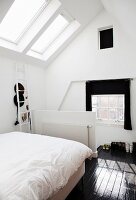 Bed with white bed linen in front of half-height balustrade wall in attic room with skylight and dark wooden floor