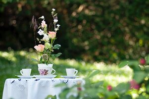 Romantic arrangement of roses in nostalgic teacup on garden table with white tablecloth and two teacups