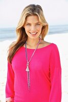 Junge, blonde Frau mit pinkem Sweatshirt und langer Kette am Strand