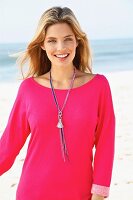 A young blonde woman on a beach wearing a pink sweatshirt and a long necklace