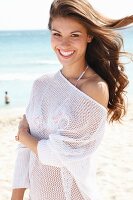 A young brunette woman on a beach wearing a white crocheted jumper