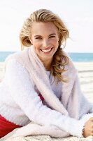 A young blonde woman by the sea wearing a light knitted jumper with another jumper over her shoulders