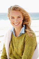 A young blonde woman by the sea wearing a knitted jumper and a denim shirt with another jumper over her shoudlers