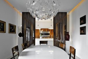Magnificent crystal lamp above white, oval dining table in front of doorway leading to modern kitchen with wood-veneer fronts
