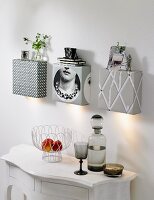 A fruit bowl and a smoky glass bottle and a matching stem glass on a white wall table with homemade wall lights covered with wallpaper that also serve as shelves hung above it