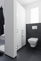 A room divider between a shower and toilet with a built in toilet paper holder and lighting with large black tiles on the floor and halfway up the wall