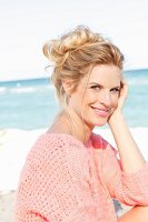 A blonde woman by the sea wearing a pink summer jumper