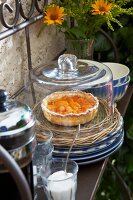 An apricot tart on a wicker plate under a glass cloche with a jug of coffee and French country house crockery on an iron shelf