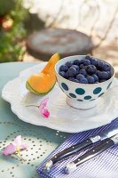 Blueberries in a spotted bowl and a wedge of honeydew melon with antique cutlery on a bistro table in a garden