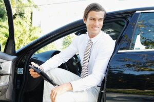 A young business man holding a tablet computer sitting in a car with the door open