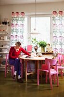 Frau auf pink lackierten Stuhl vor rundem Tisch am Fenster, bodenlange Vorhänge mit Blumenmuster