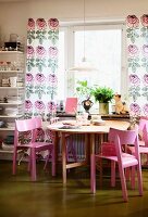 Pink wooden chairs around round table below window with floor-length, floral curtains