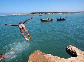 Junge macht Kopfsprung vom Felsen ins Meer, im Hintergrund Fischerboote, Larache, Marokko