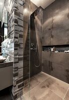 An open rain shower with dark tiles and a glass partition wall with limestone relief tiles behind the sink