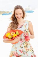 Junge Frau im Sommerkleid hält mit Obst gefüllten Hut und bietet einen Apfel an