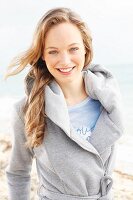 A young blonde woman by the sea wearing a grey sweatshirt coat