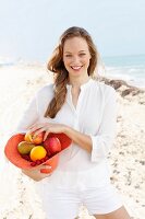 Junge Frau in weisser Bluse und Shorts hält mit Obst gefüllten Hut am Meer