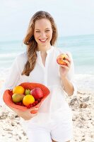 A young woman by the sea wearing a white blouse and shorts holding a hat filled with fruit