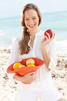 A young woman by the sea wearing a white blouse holding a hat of fruit