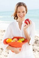 A young woman by the sea wearing a white blouse and holding a hat of fruit