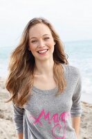 A young blonde woman by the sea wearing a grey jumper with pink writing