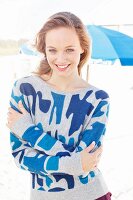 A young blonde woman on a beach wearing a blue patterned jumper