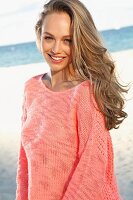 A young blonde woman by the sea wearing a pink summer jumper
