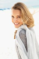 A blonde woman on a beach wearing a grey cardigan and a scarf