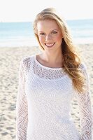 A young blonde woman on a beach wearing a white top and a transparent knitted jumper