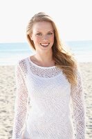 A young blonde woman on a beach wearing a white top and a transparent knitted jumper