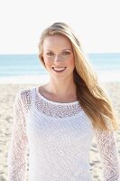 A young blonde woman on a beach wearing a white top and a transparent knitted jumper
