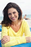 A brunette woman on a beach with her arms folded wearing a yellow knitted jumper