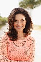 A brunette woman on a beach wearing a salmon-coloured top and an openwork jumper