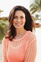 A brunette woman on a beach wearing a salmon-coloured top and an openwork jumper