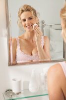 A blonde woman in front of a mirror cleaning her face with a cotton wool pad
