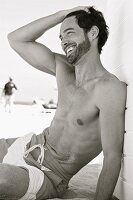 A laughing young man sitting on a beach wearing swimming shorts (black-and-white shot)