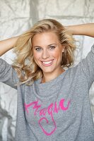 A blonde woman with her arms behind her head wearing a grey jumper with the word 'Magic' in pink