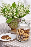 Plätzchen im Glasbehälter und auf Teller vor Maiglöckchenstrauss in Vase, Tischdecke mit weiss braunem Punktemuster