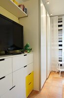 A white bedroom with yellow accents and a television on top of a chest of drawers next to a wardrobe