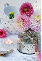 Pink dahlias in mercury glass vase on lace doily with teacups in background