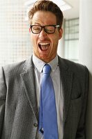 A man wearing glasses, a grey blazer and a tiny making a face: winking