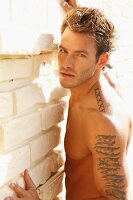 A young, topless, tattooed man leaning against a wall