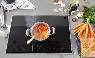 A carrot medley in a steaming pot on an induction stove surrounded by ingredients and cooking utensils
