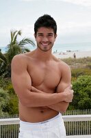 A young, topless man with his arms folding standing on a terrace by the sea