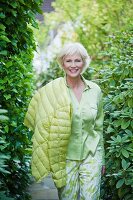 An older woman wearing a light green blouse, green-and-white trousers and a quilted jacket in a garden