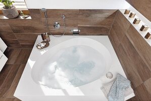 Bubbles in a droplet-shaped bath set in a white mineral-cast block with wooden-style tiles on the walls and floor