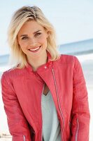 A blonde woman by the sea wearing a light jumper and a salmon pink leather jacket