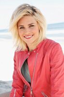 A blonde woman by the sea wearing a light jumper and a salmon pink leather jacket
