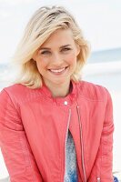 A blonde woman by the sea wearing a blue jumper and a salmon coloured leather biker jacket